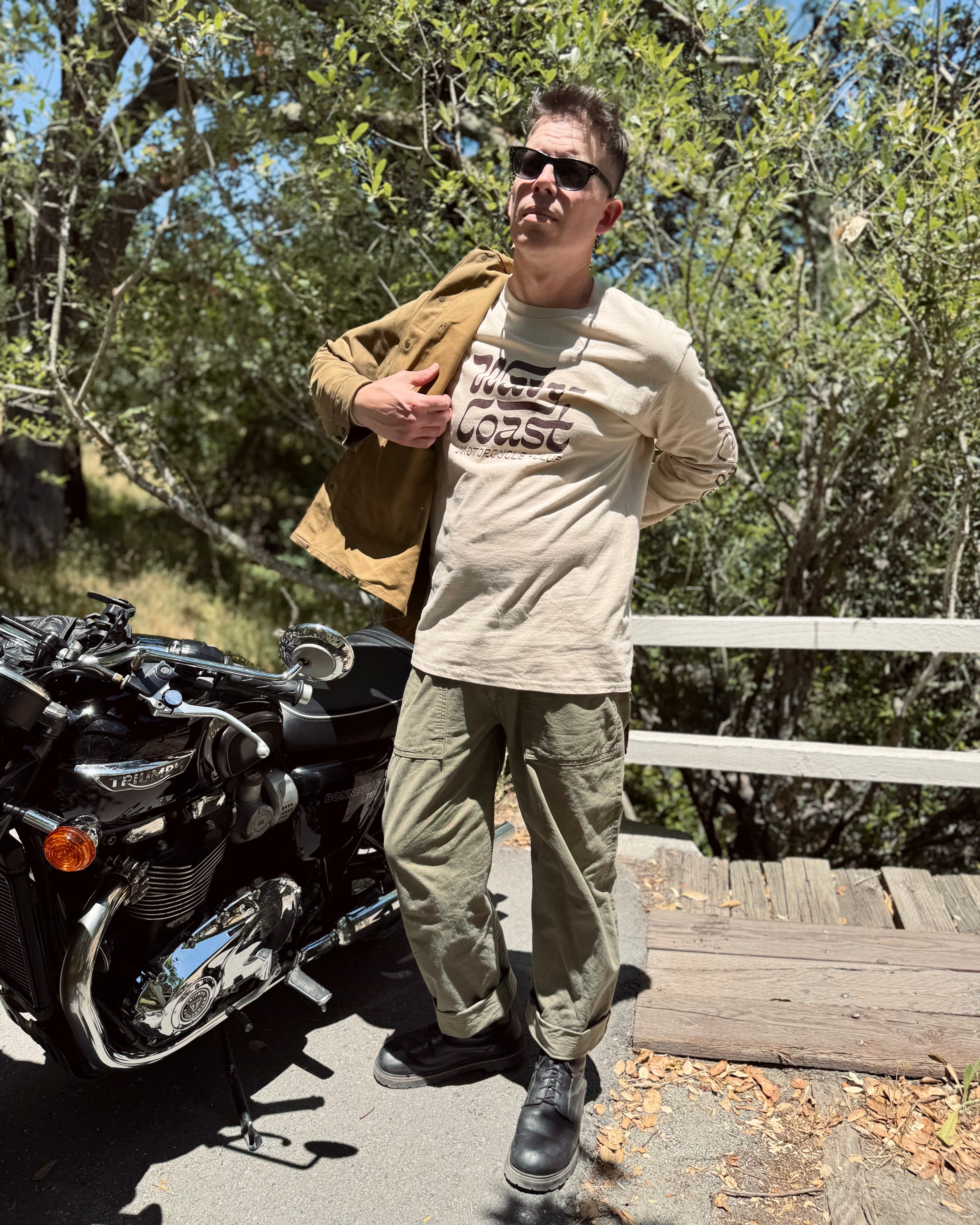 A man wearing a beige graphic t-shirt, olive green cargo pants, and black boots stands next to a black motorcycle outdoors on a wooden bridge with trees in the background.