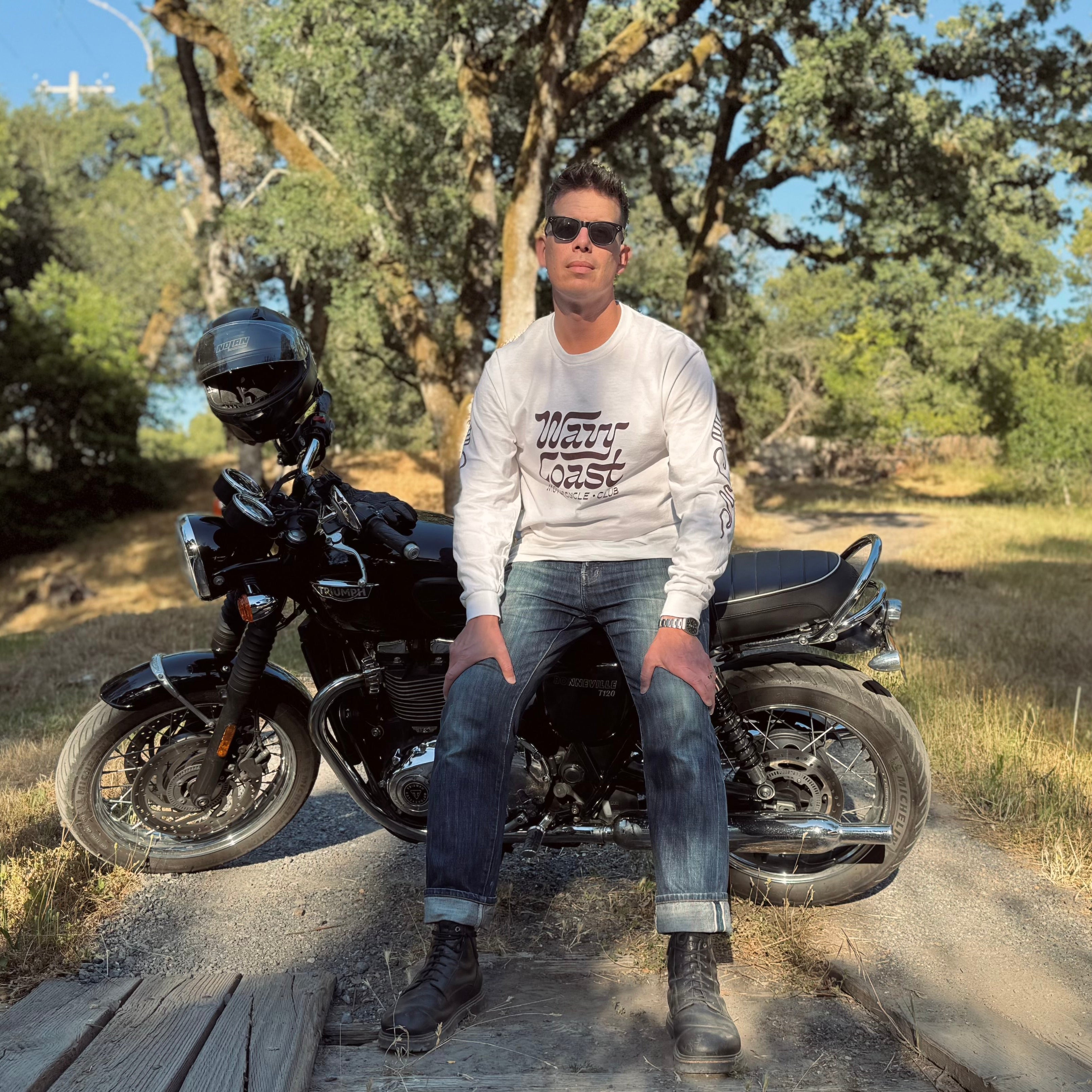 A man wearing a white graphic long-sleeve shirt, blue jeans, and black boots sits on a black cruiser motorcycle on a wooden bridge outdoors. Trees and a grassy field are visible in the background.